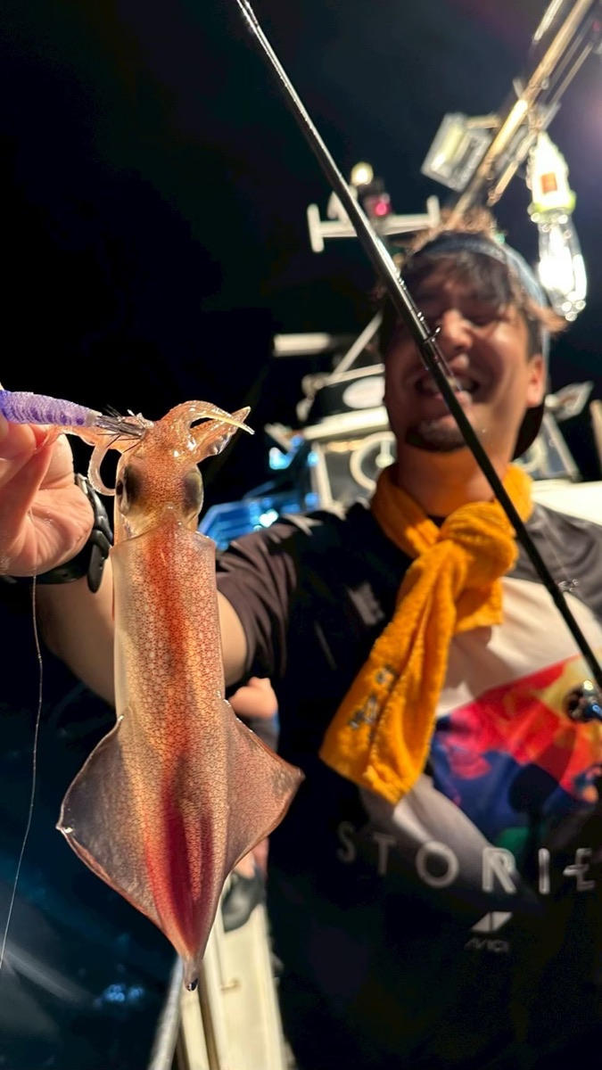 夜の船上で、男性が釣り上げたマイカを手前に掲げて笑っている